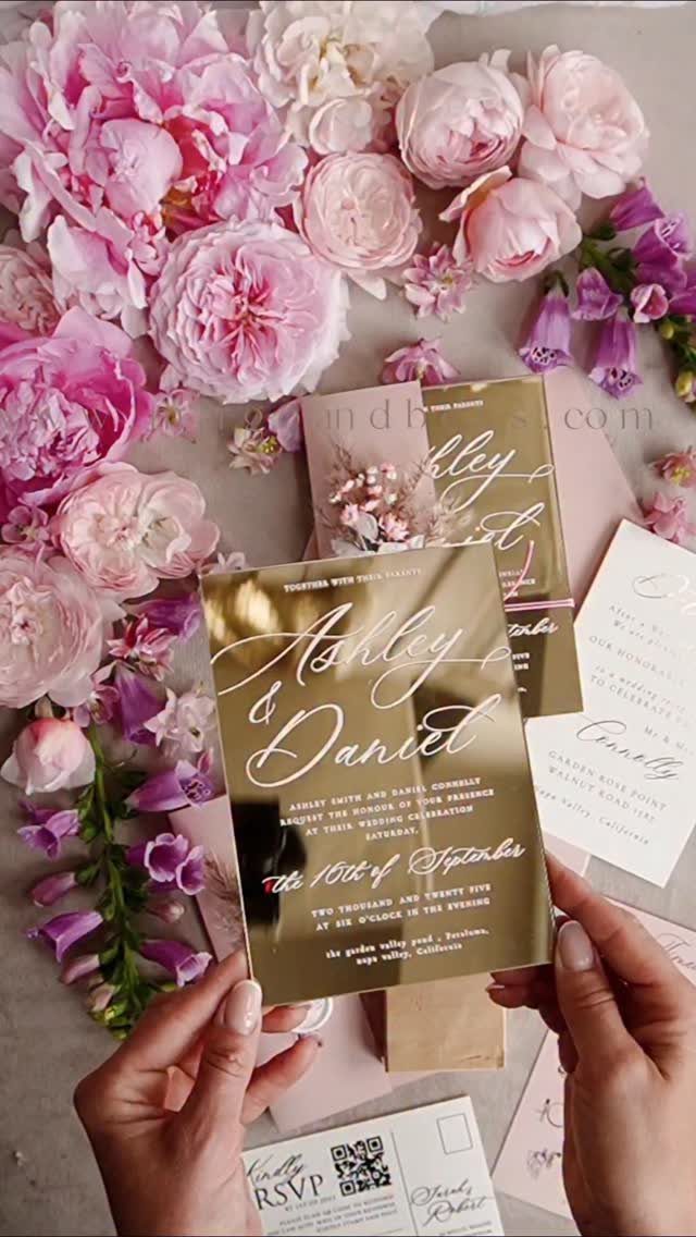 Rose gold metallic wedding invitation held by hands surrounded by pink peonies