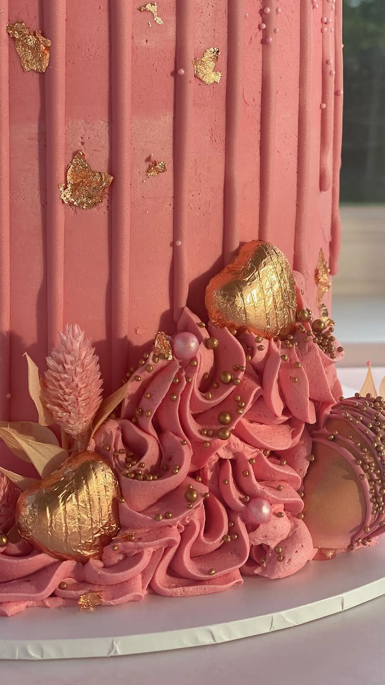Pink frosted cake decorated with rose gold hearts, gold leaf, and metallic pearls.