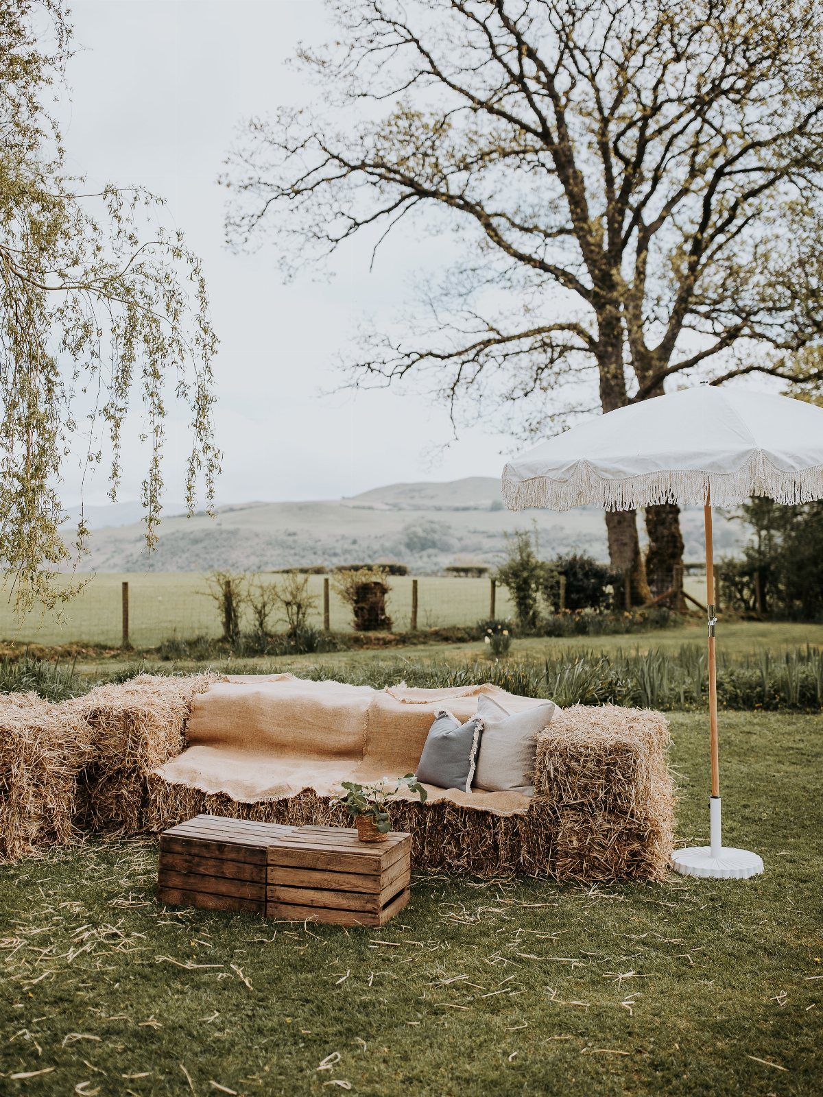 Hay bale seating with tan fabric and pillows under a white fringed umbrella.