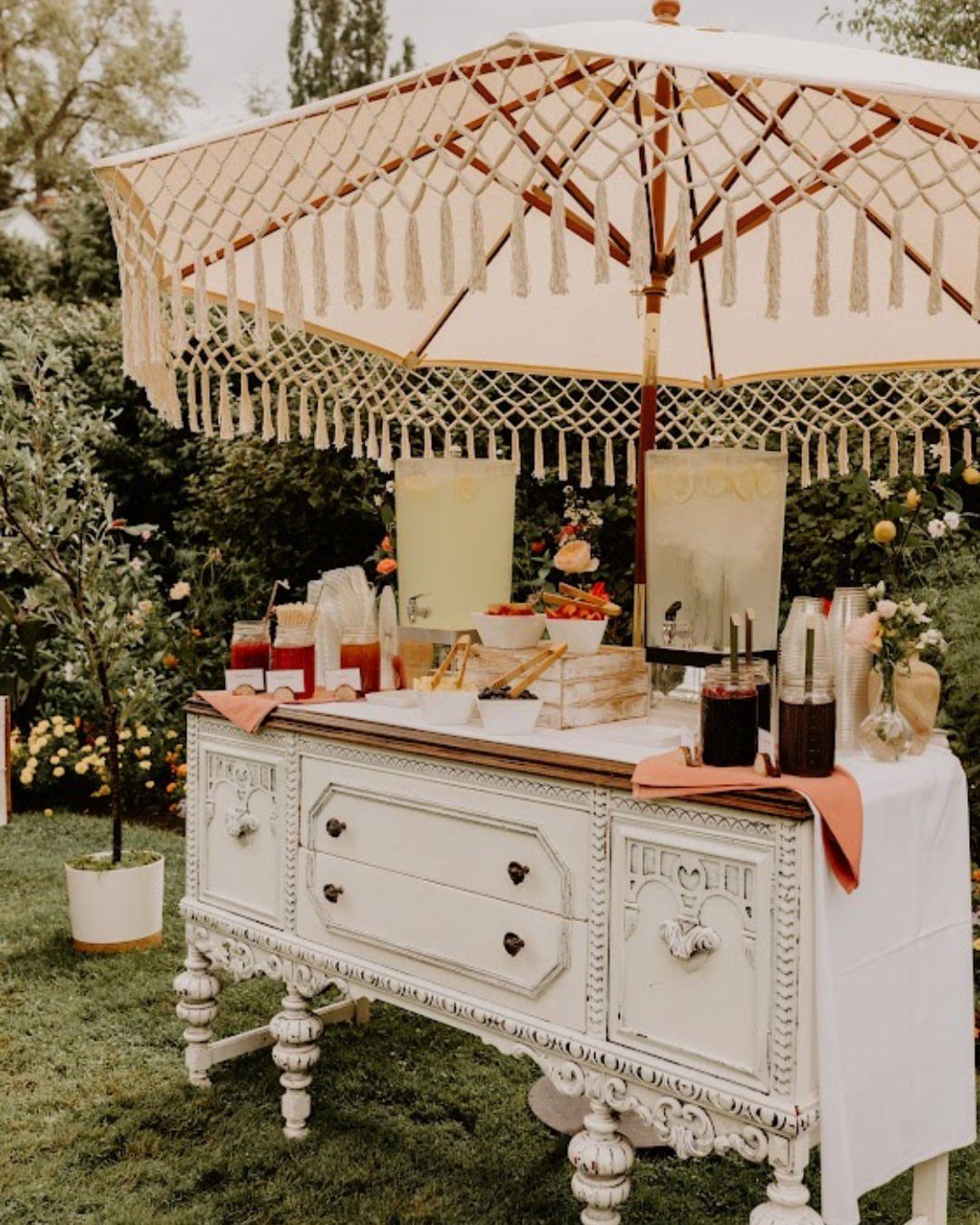 Vintage white sideboard drink station with colorful beverages under a fringed cream umbrella.