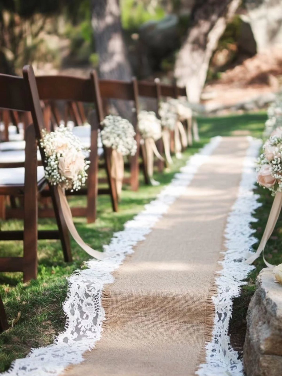 Burlap aisle runner with lace trim between wooden chairs with white floral bouquets.