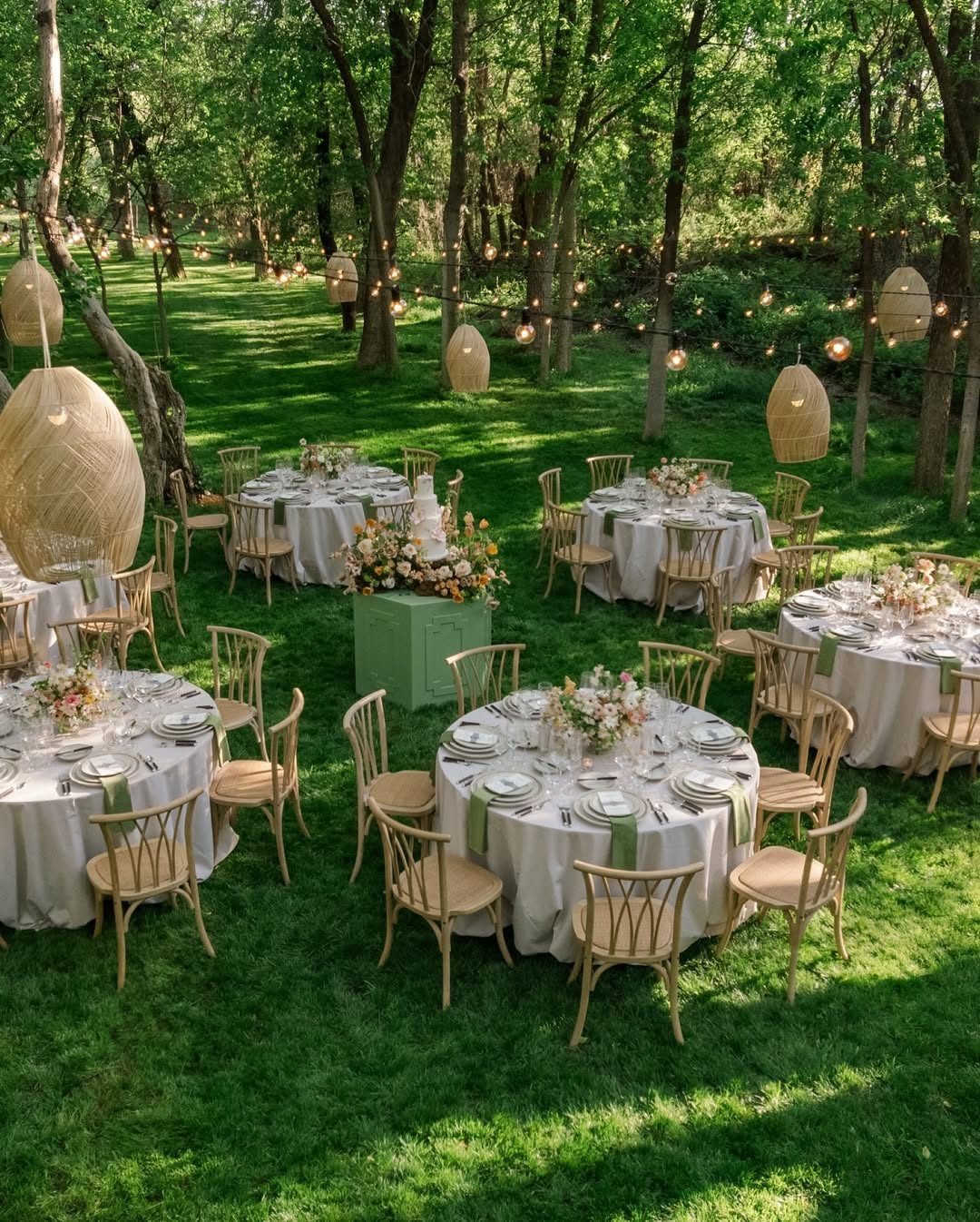 Woven lanterns and string lights hanging over white dinner tables in a forest