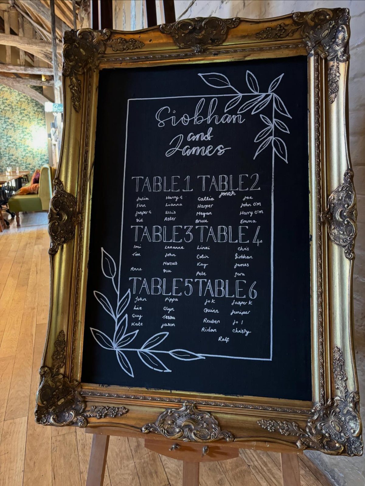 Ornate gold framed chalkboard displaying a wedding seating chart on a wooden easel.