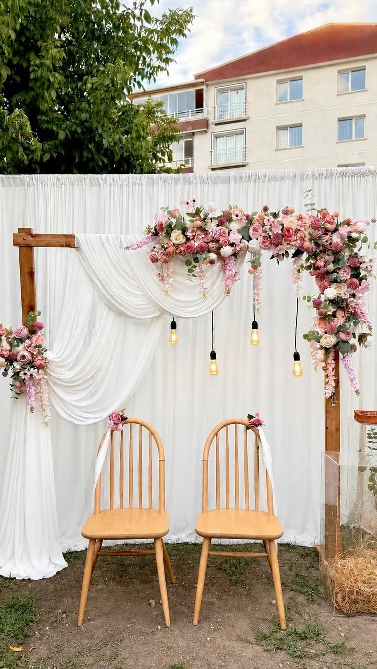 Wooden wedding arch draped in white fabric and pink flowers with two wooden chairs.