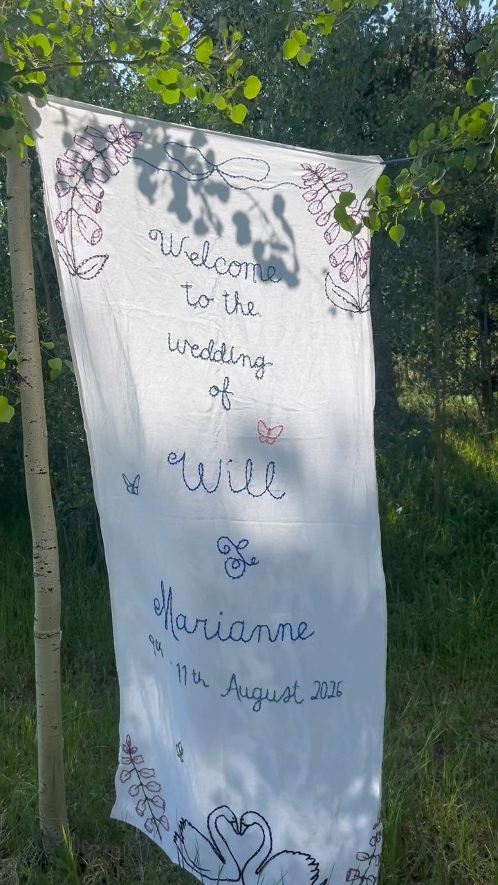 White embroidered wedding welcome banner hanging from a tree in a green forest