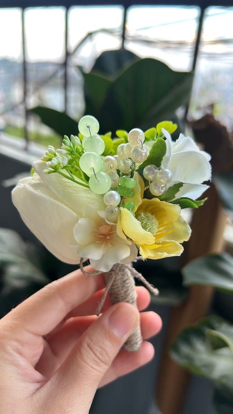 Floral boutonniere with white and yellow flowers and sage green berries held by hand.