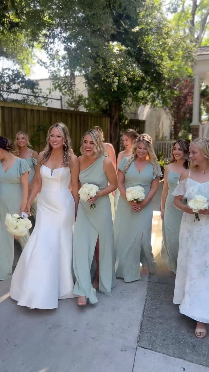 Bride in white gown walking with bridesmaids wearing sage green dresses and bouquets