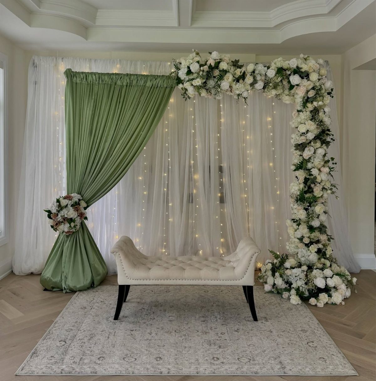 Sage green draped curtain and white floral arch backdrop with a white chaise lounge.