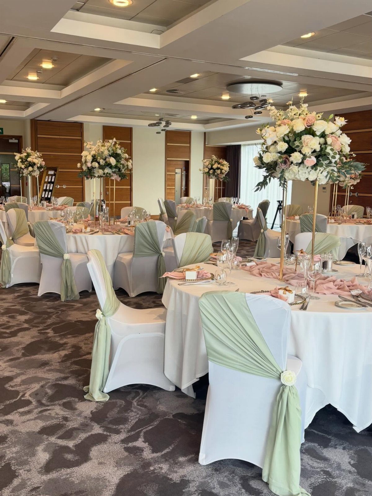 Wedding reception tables with white linens, sage green chair sashes, and floral centerpieces.
