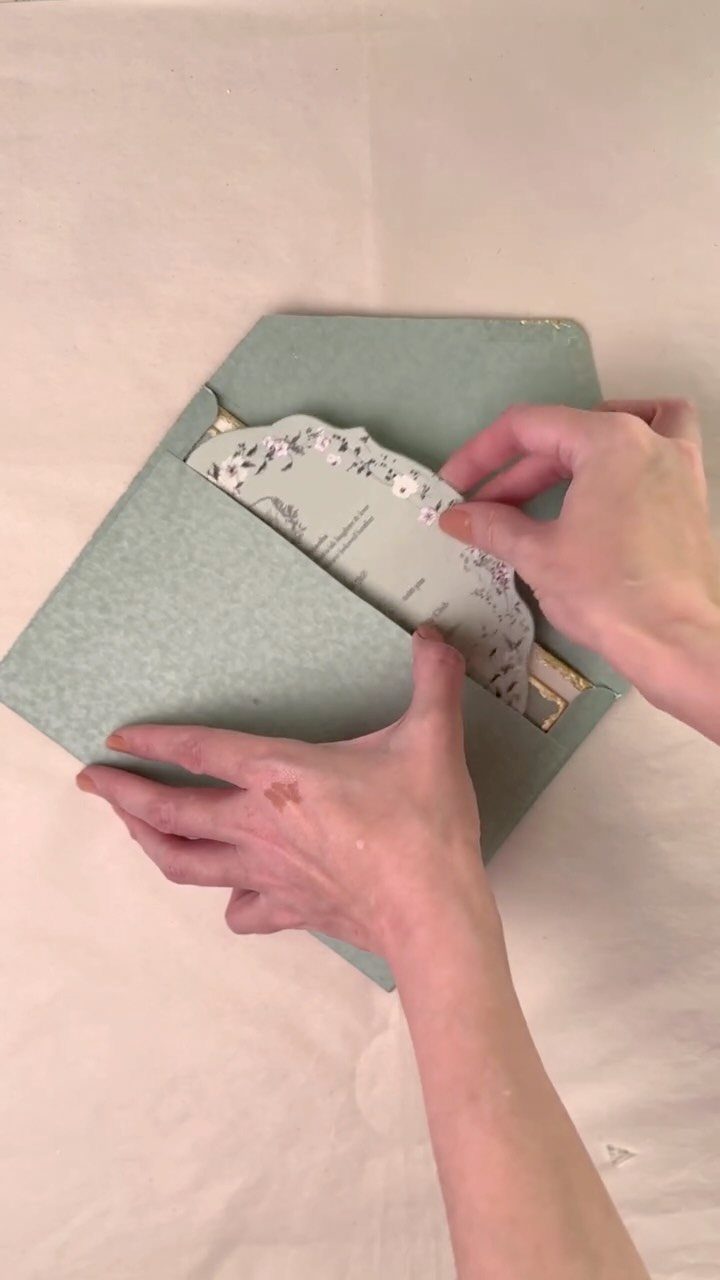 Hands removing a floral wedding invitation from a sage green textured envelope.