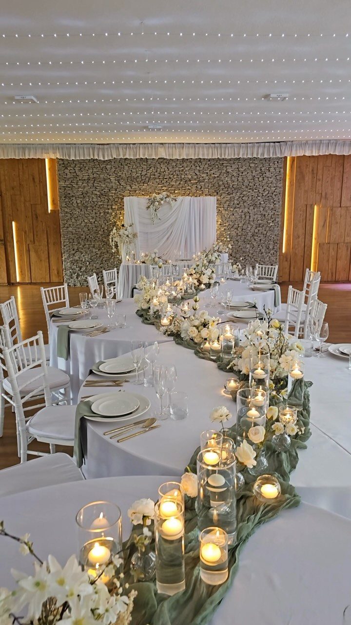 Wedding reception table with sage green runners, white flowers, and glowing cylinder candles.