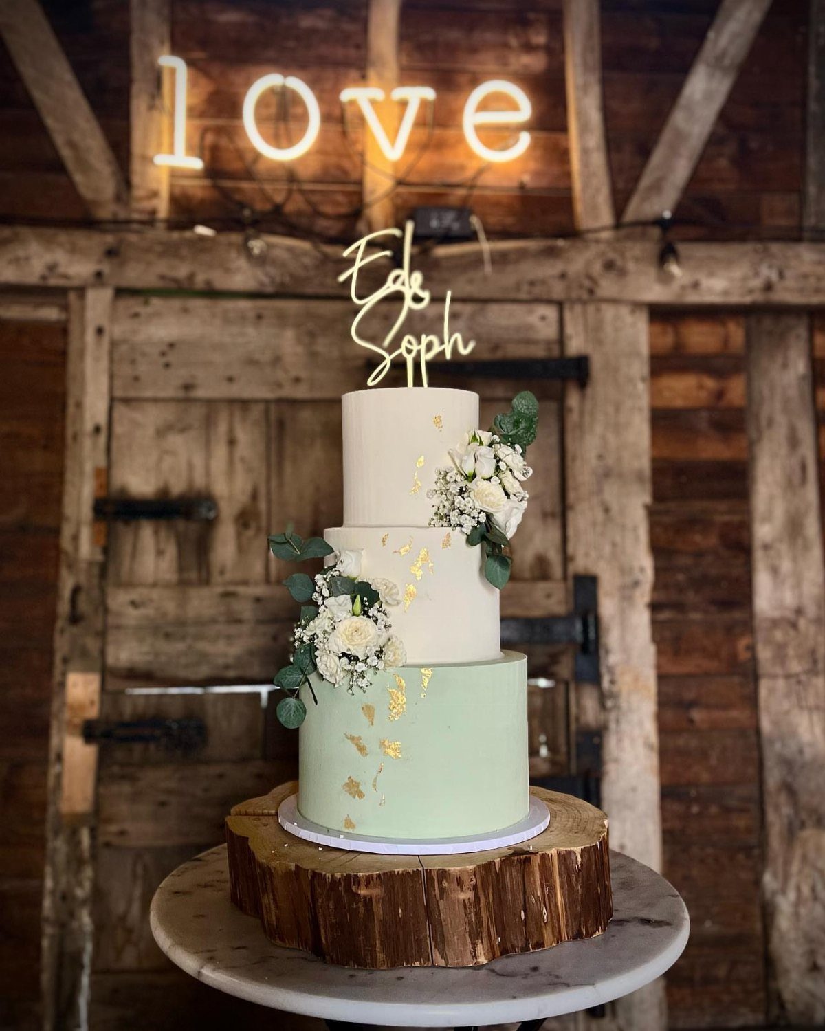 Three-tier wedding cake with sage green base and white flowers on wood slice.