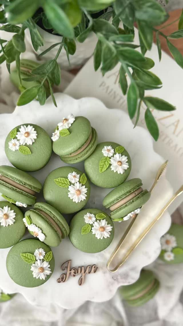 Sage green macarons with white frosting flowers on a white scalloped plate.