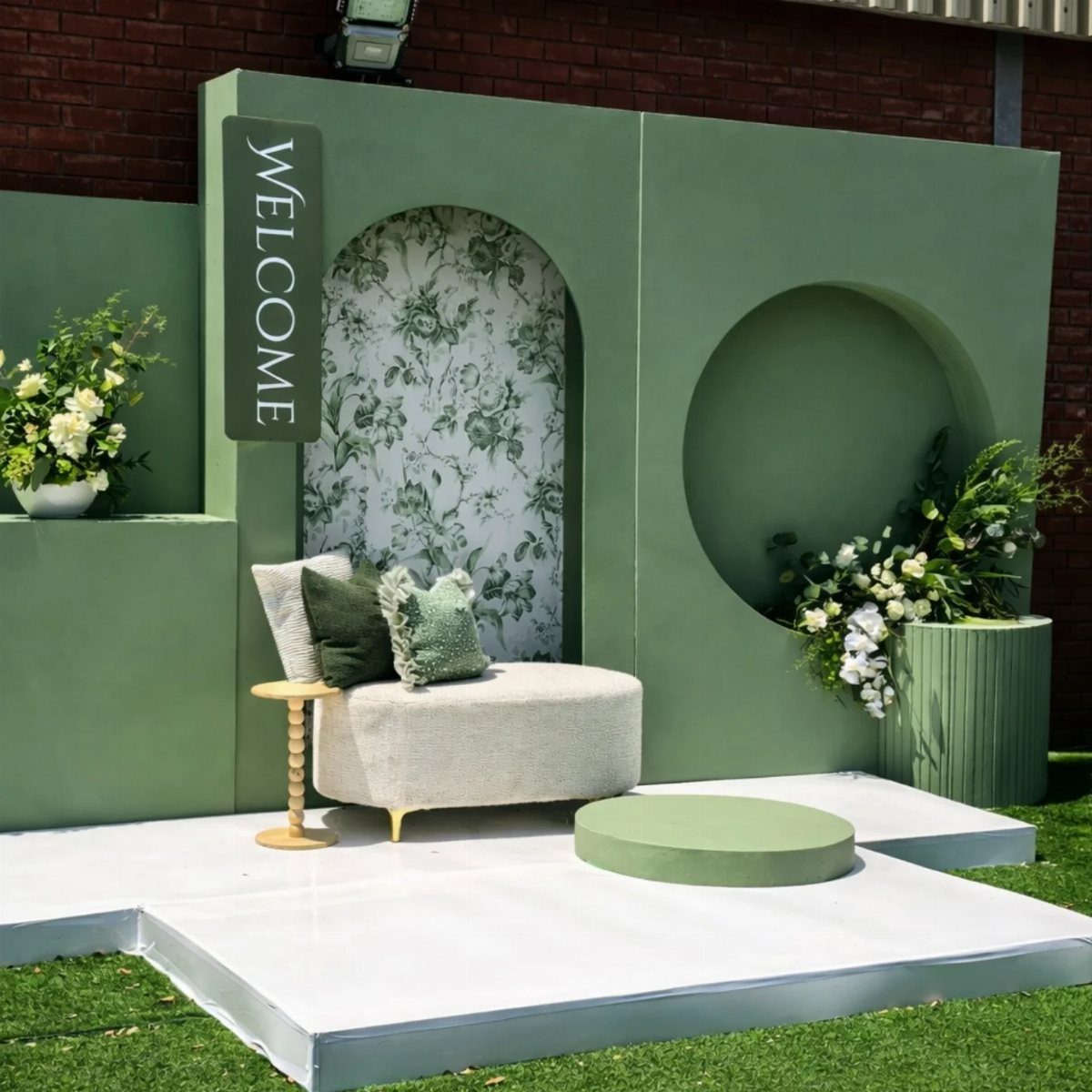 Sage green event backdrop with a white lounge chair and floral accents outdoors.