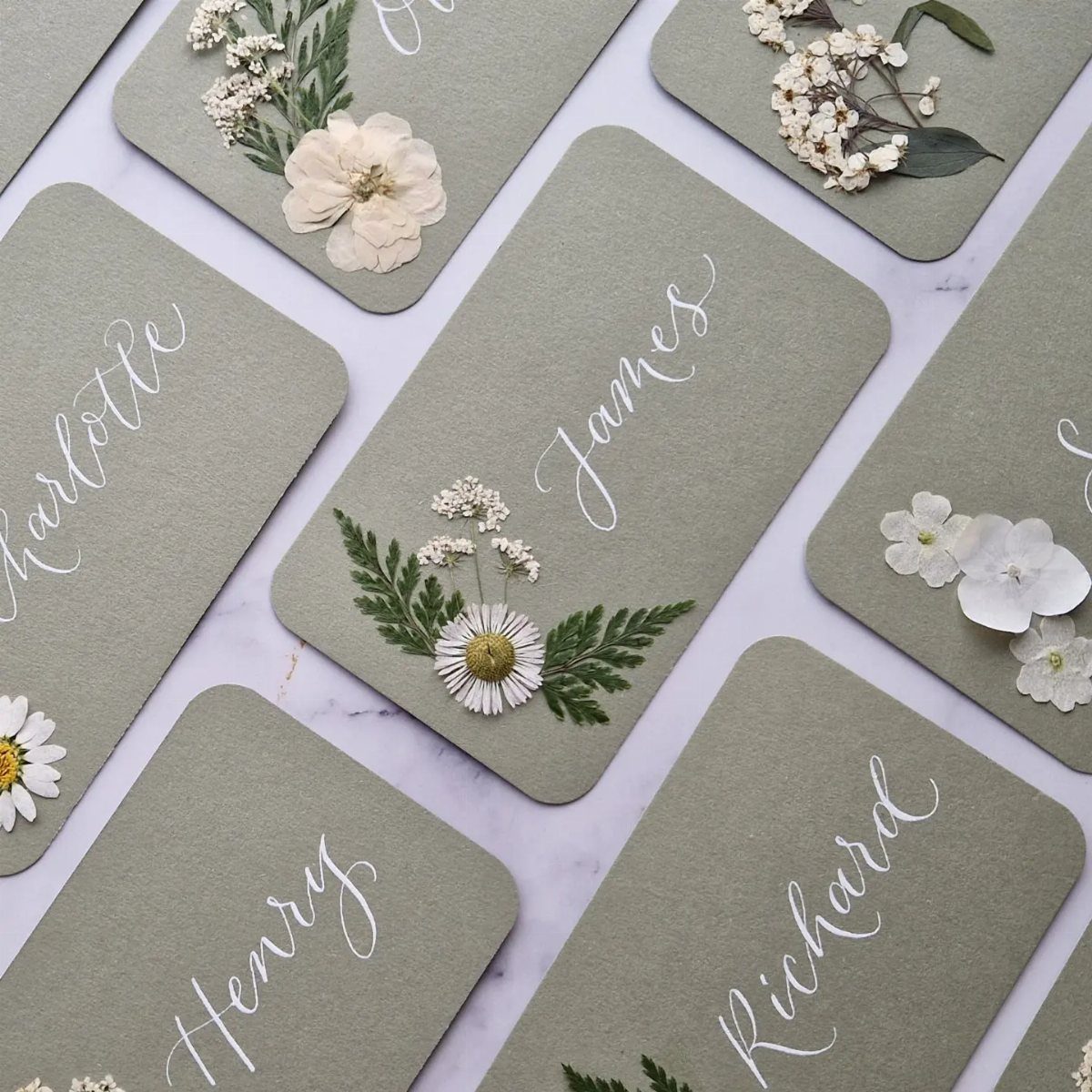 Sage green place cards with white cursive names and dried floral decorations on marble.
