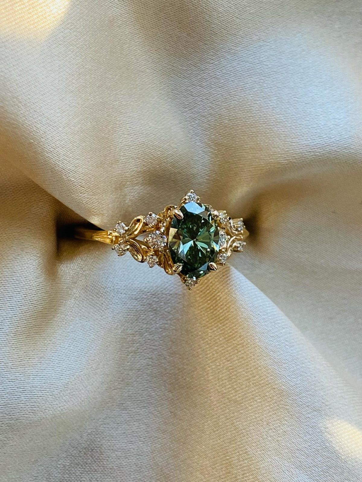 Gold ring with a sage green gemstone resting on cream satin fabric