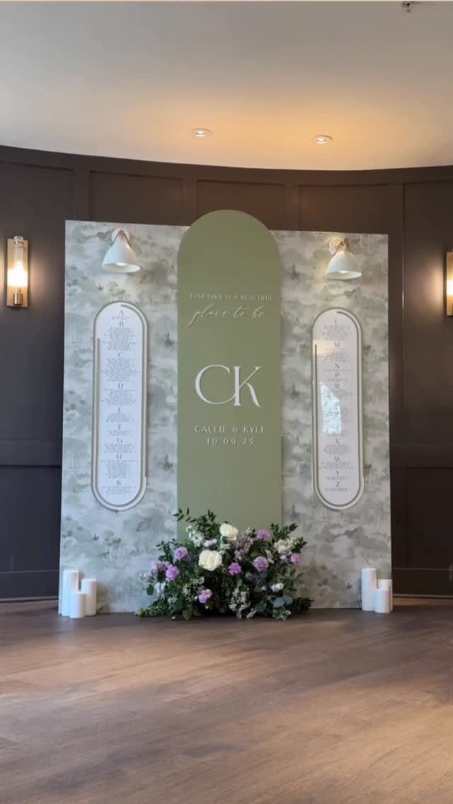 Sage green arched wedding sign with seating charts and purple floral arrangements.