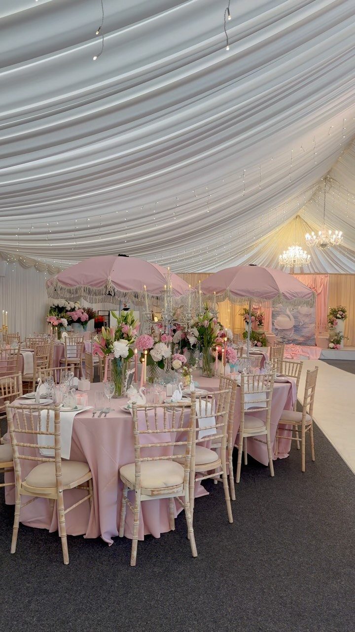 Round wedding table with pink linens, gold chairs, and pink umbrellas in a marquee.