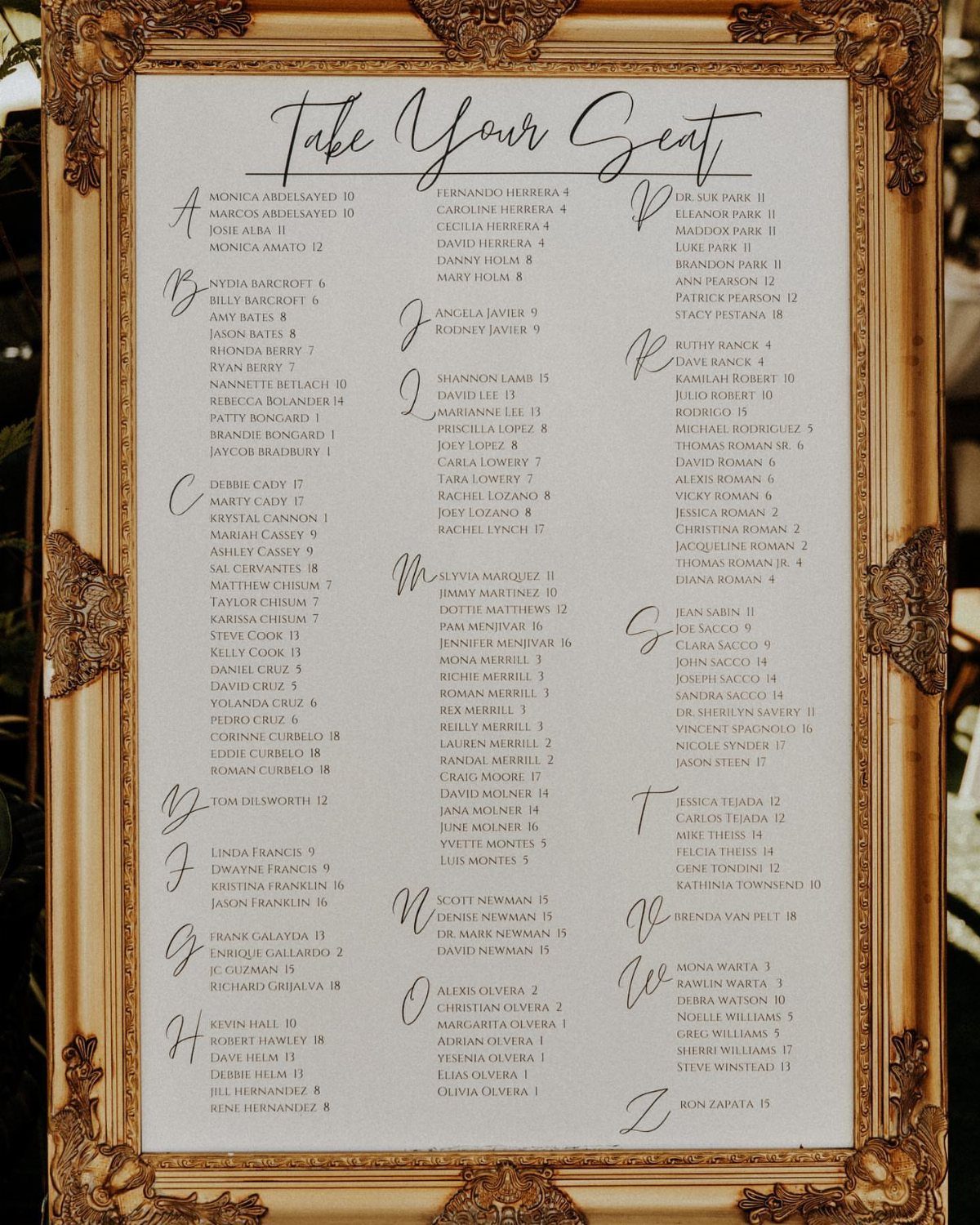 Alphabetical wedding seating chart in an ornate gold frame with white background.