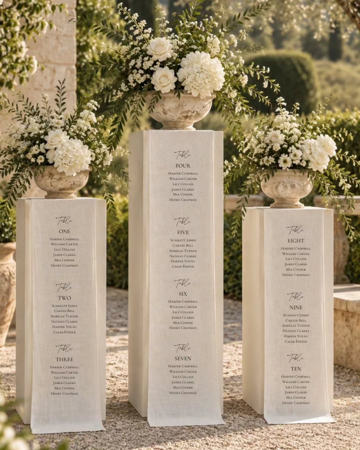 Three white column pedestals with floral urns serving as a wedding seating chart.