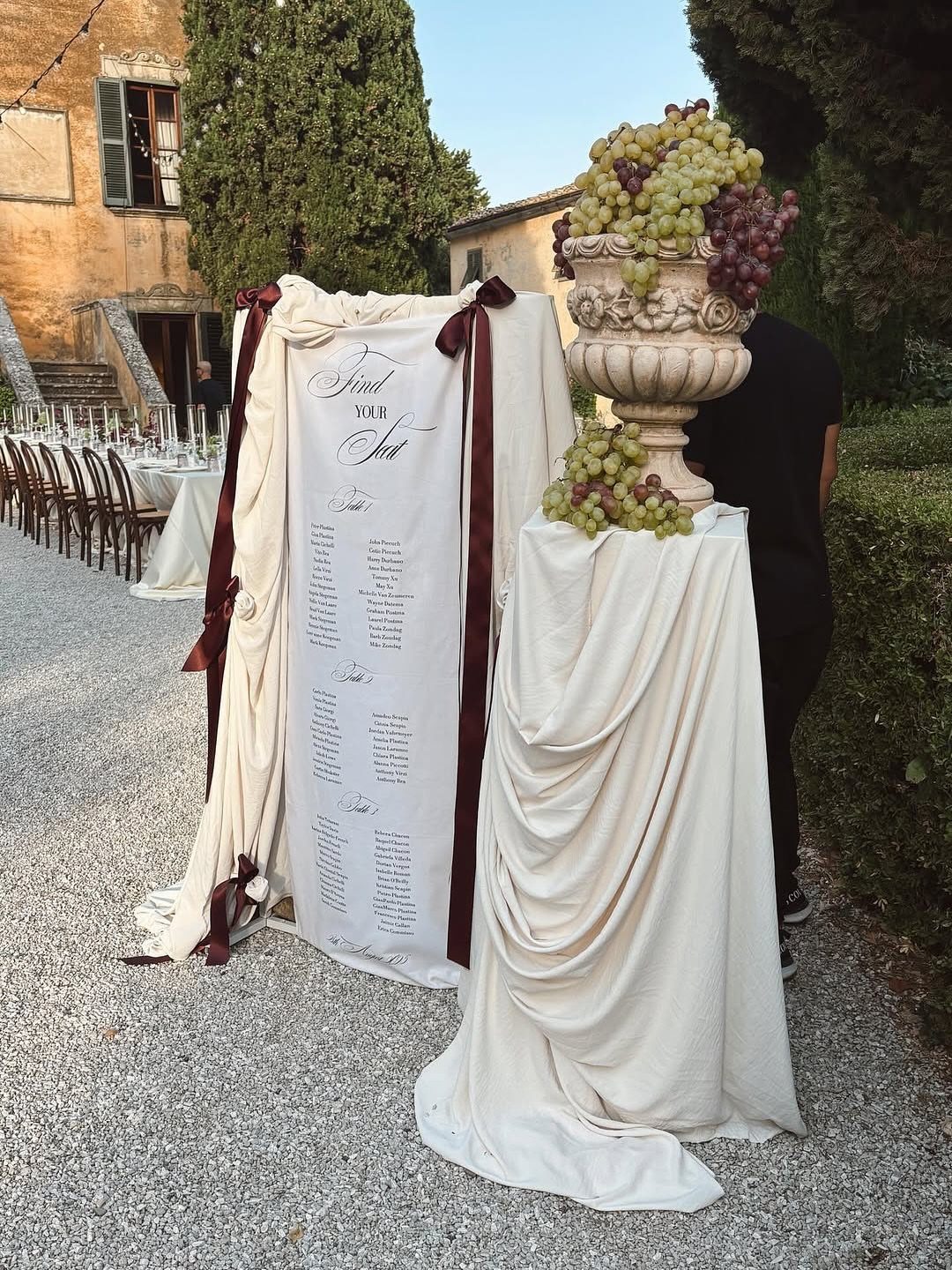 White fabric seating chart with burgundy ribbons next to a grape-filled stone urn.