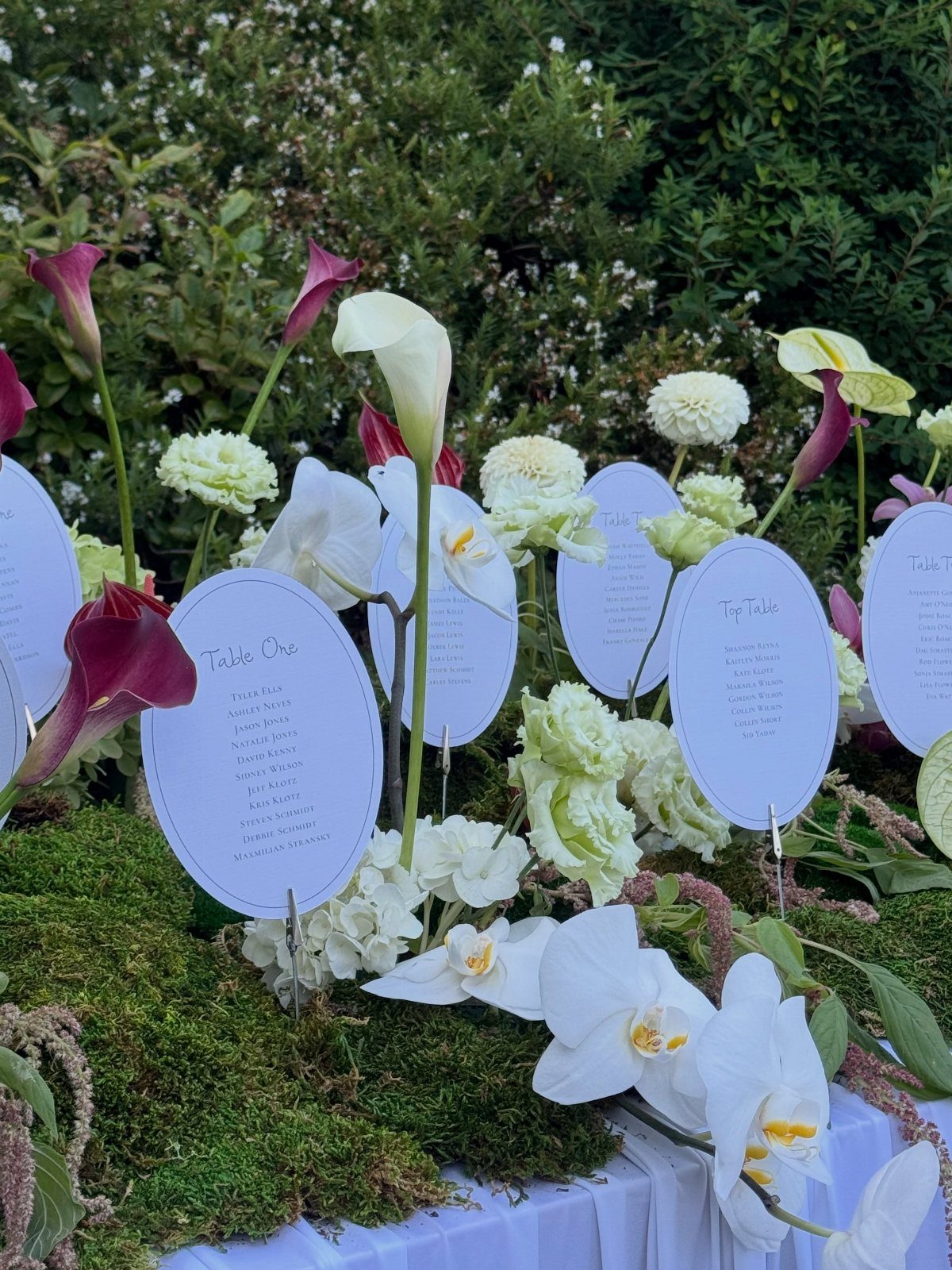 White oval table cards nestled in white orchids and calla lilies on green moss.