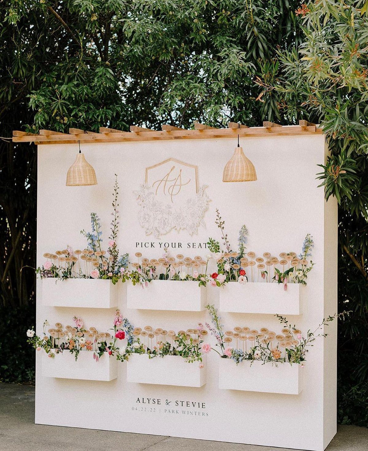 White seating chart wall with built-in flower planters and a wooden trellis top.