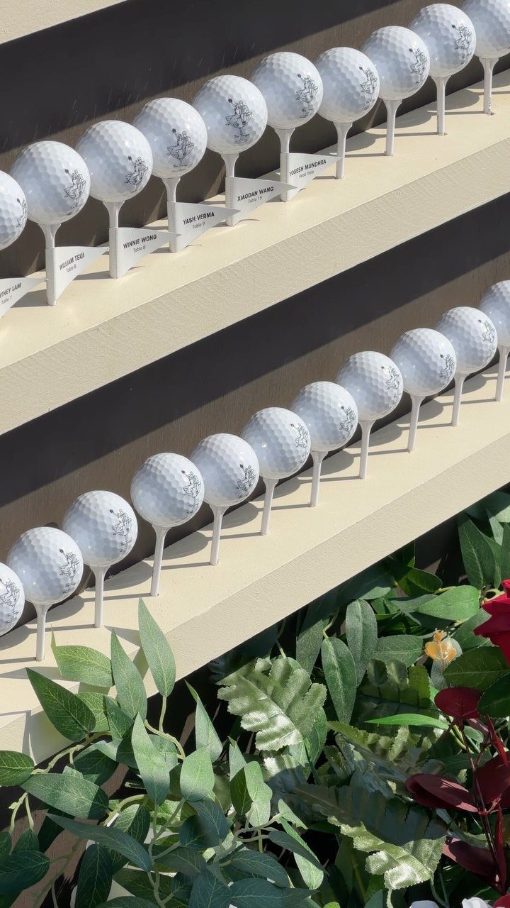 Golf balls on tees with name tags arranged on tiered shelving with greenery.