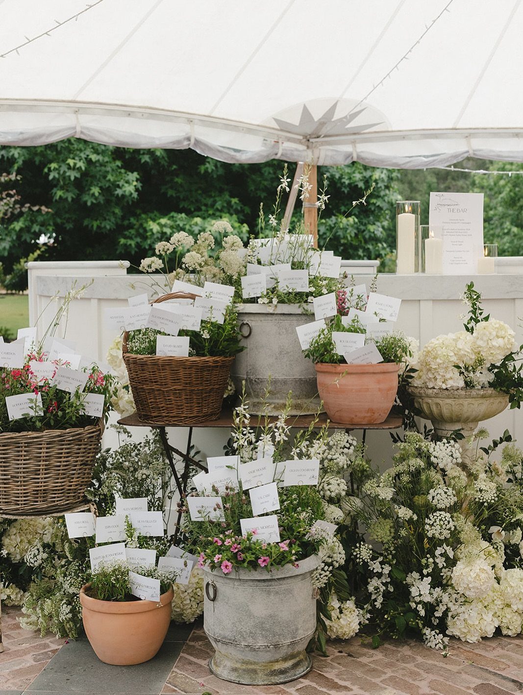 Potted plants and wicker baskets with white escort cards under a white tent.
