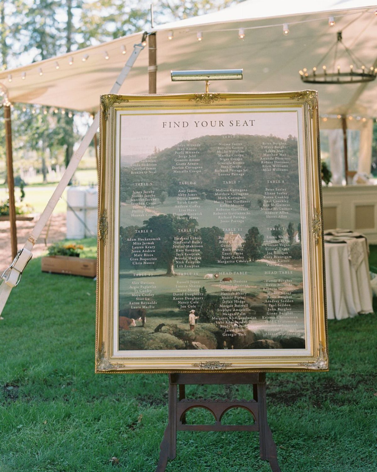 Gold vintage frame seating chart on a wooden easel under a white tent.