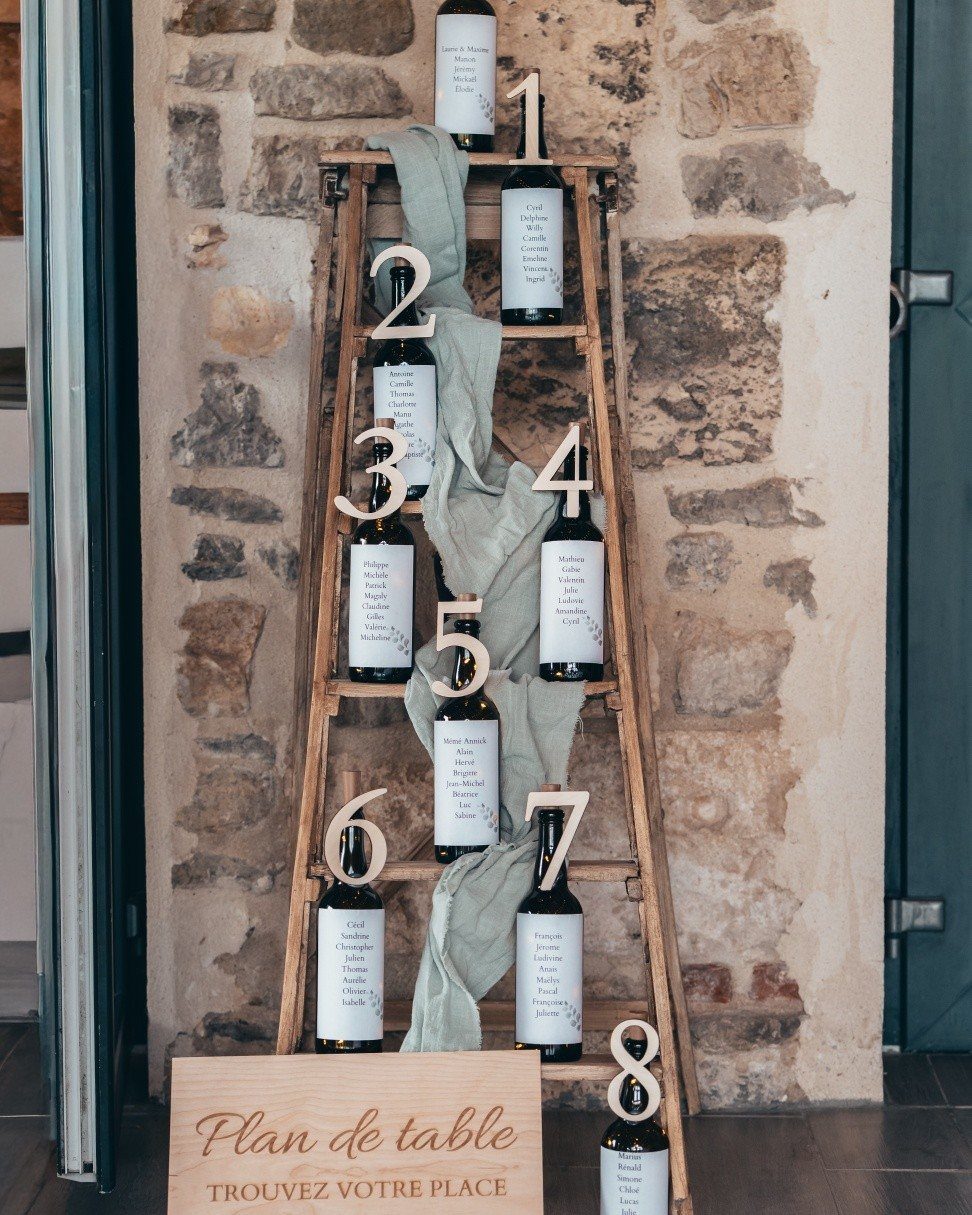 Wooden ladder seating plan with wine bottles and numbers against a stone wall.