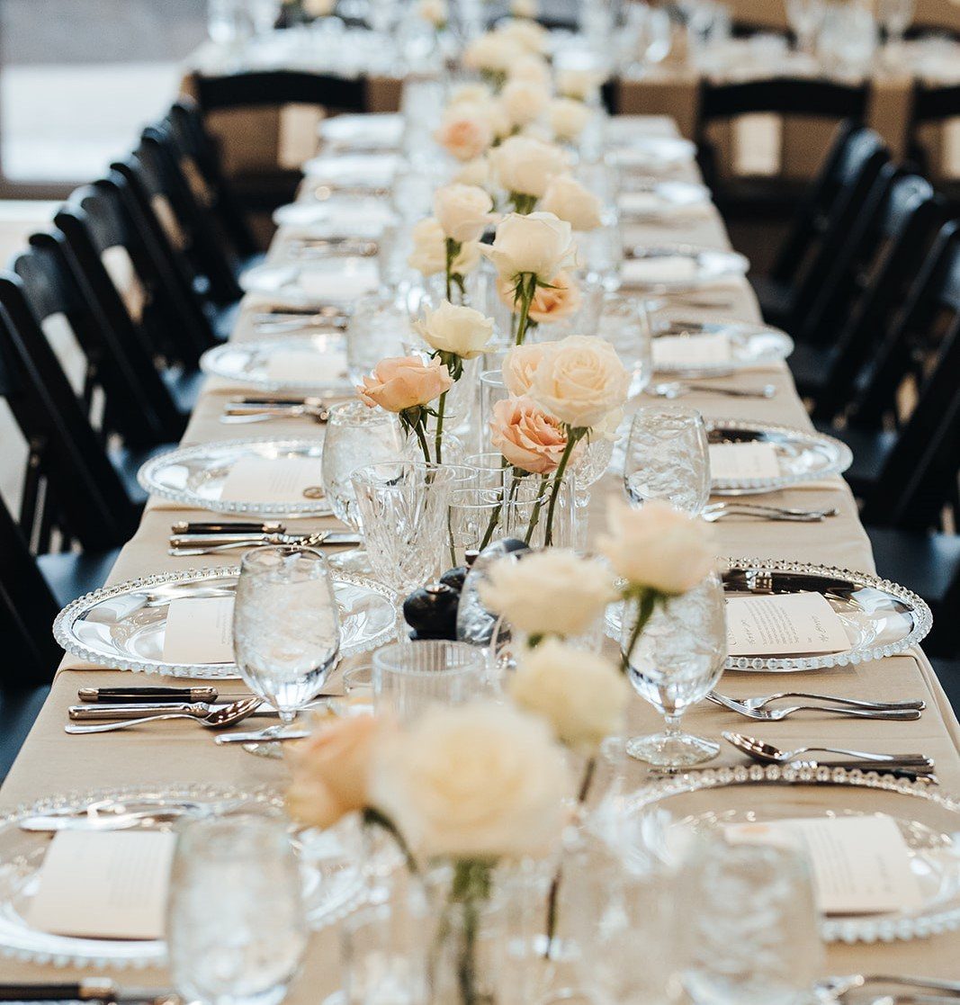 Single pale peach roses in small clear glass vases along a wedding table.