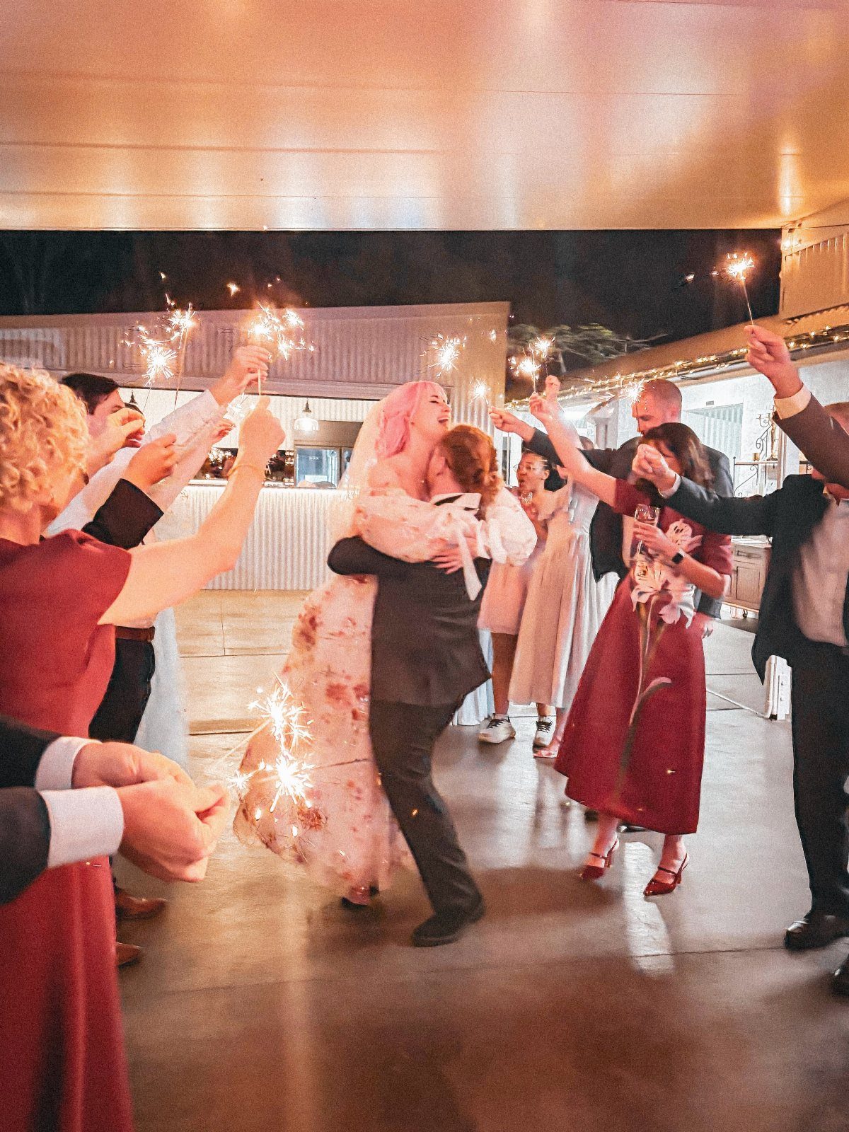 Bride and groom embracing while guests hold lit sparklers during a wedding celebration.