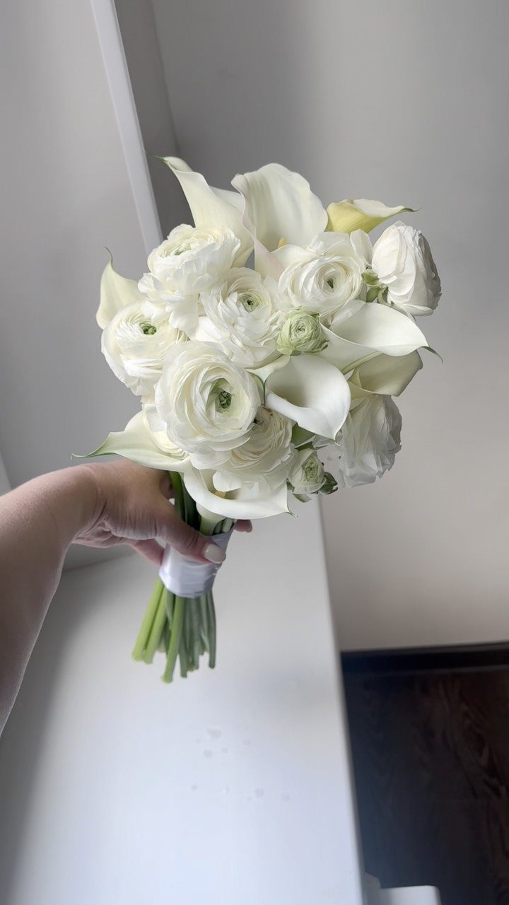 White bridal bouquet with calla lilies and ranunculus held by a hand.