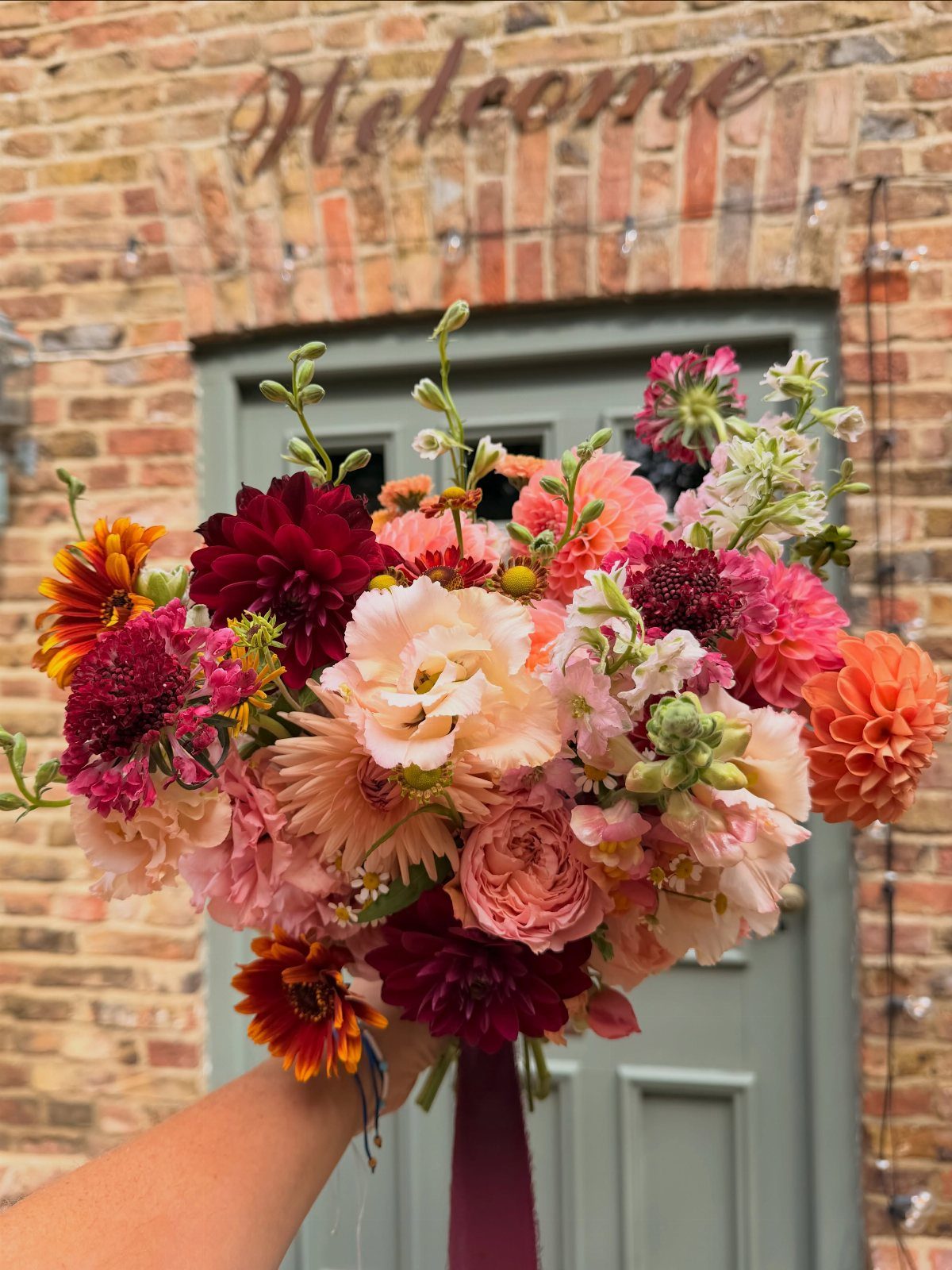 Vibrant wedding bouquet with bold pink, coral, and burgundy dahlias against brick wall.