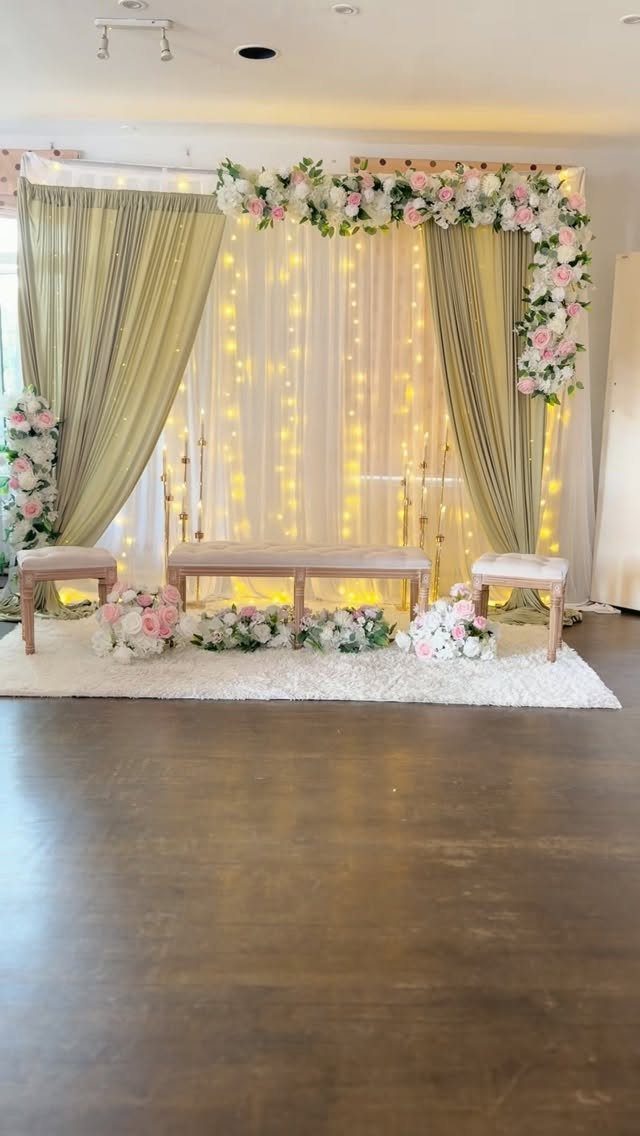 Sage green curtains and white floral arch backdrop with fairy lights and wooden bench.