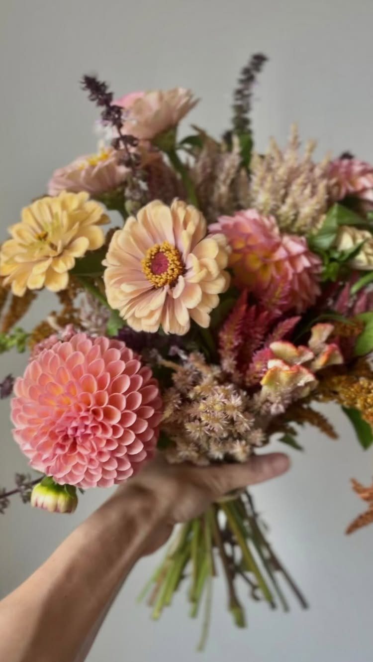 Floral bouquet with pink, peach, and yellow flowers held by a hand.