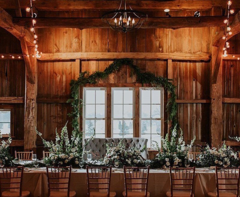 Rustic wooden barn interior with wedding table, white flowers, and greenery arch.