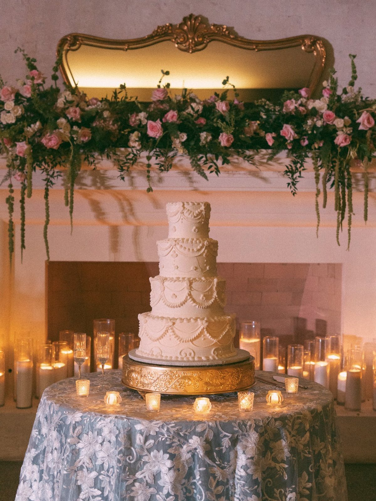 Four-tier white wedding cake on a blue floral tablecloth with floral mantel backdrop.