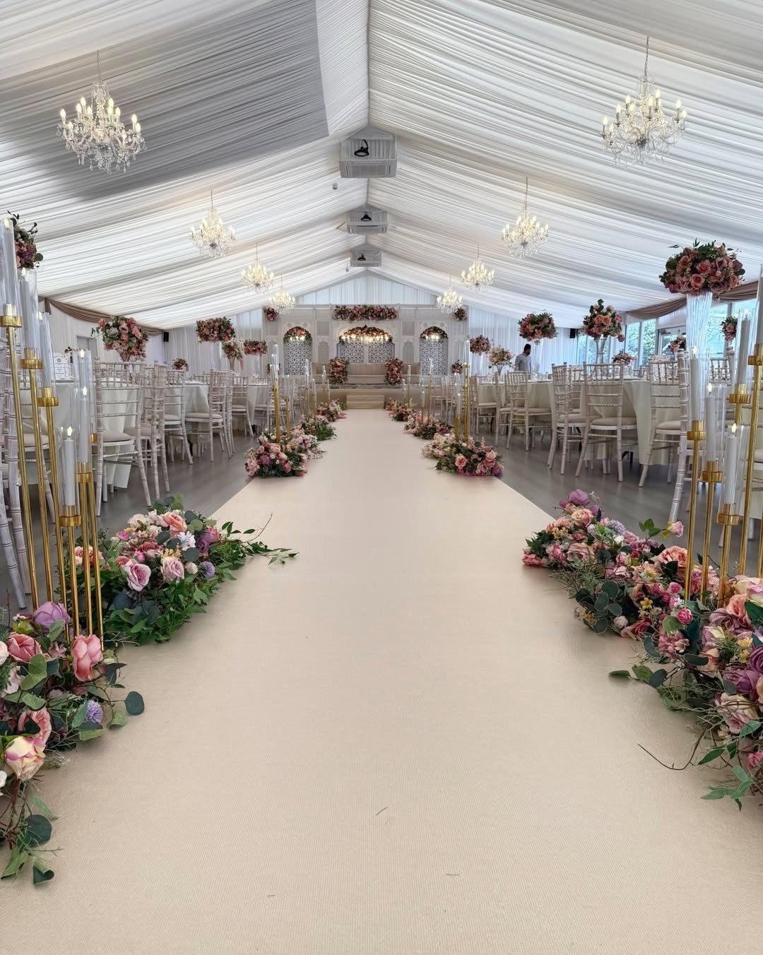 White wedding tent with crystal chandeliers and pink rose arrangements lining a cream aisle.