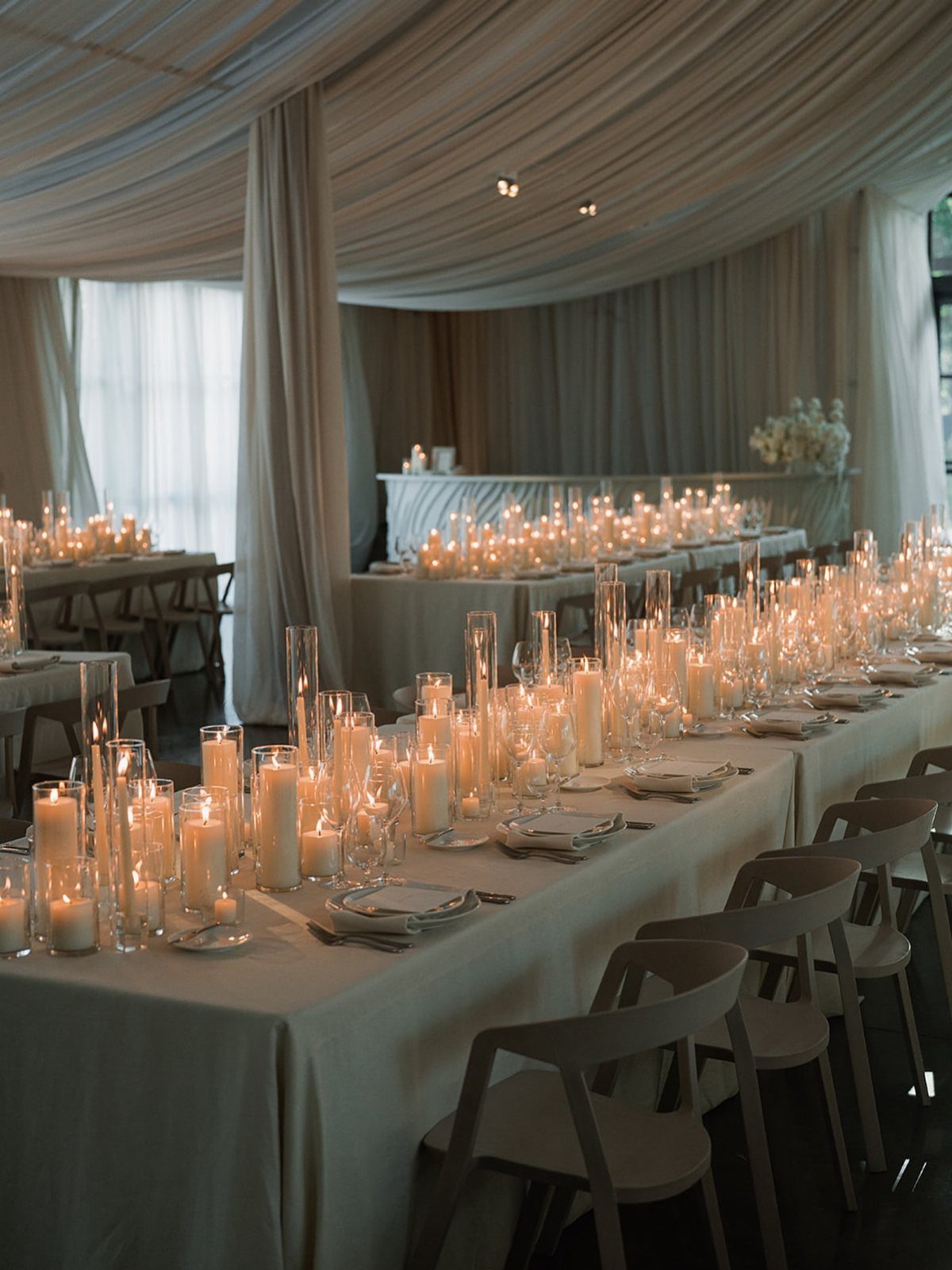 White pillar candles in glass cylinders lining long white wedding reception tables.