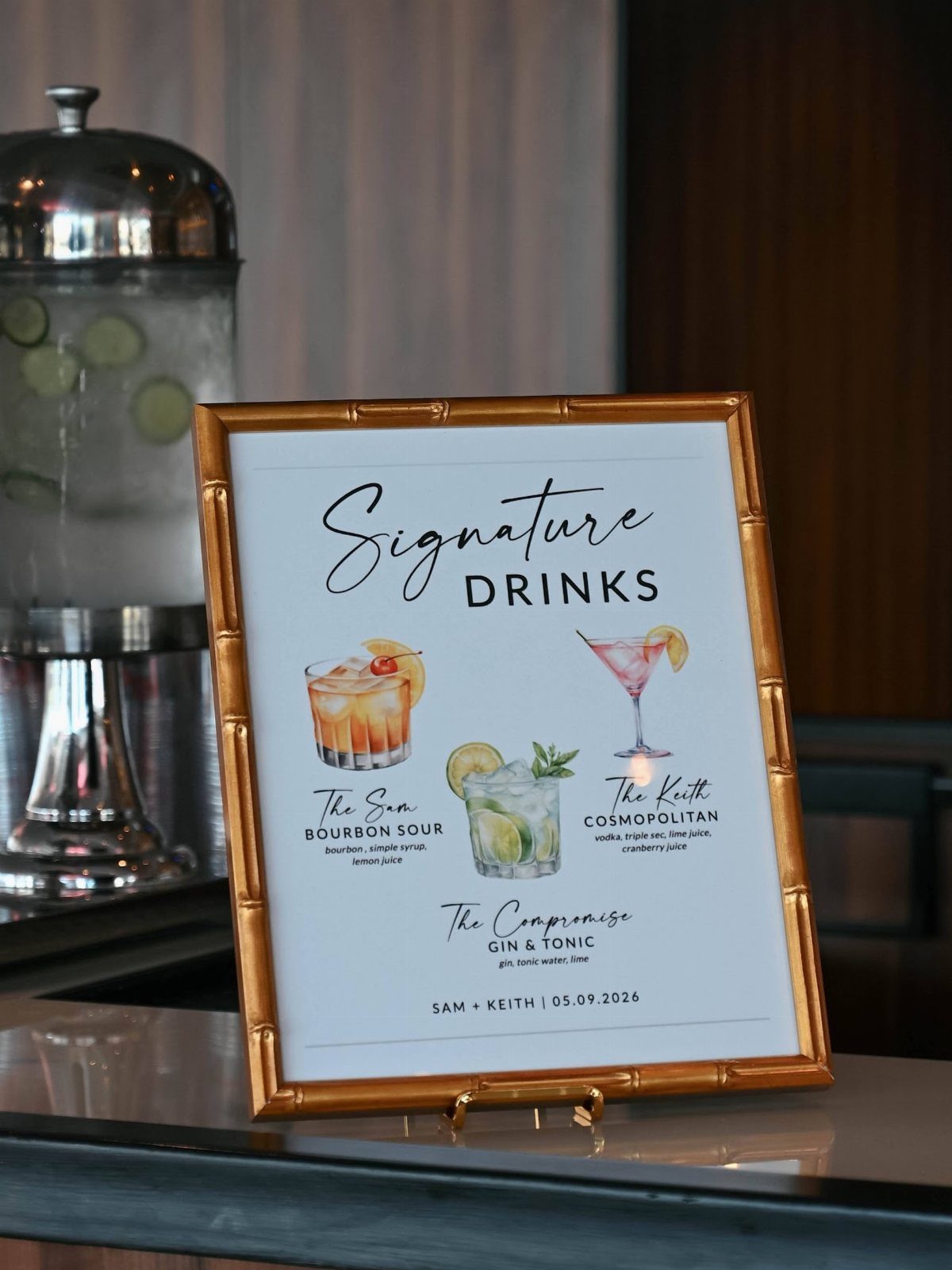 Gold-framed signature drinks menu with cocktail illustrations on a wedding reception counter