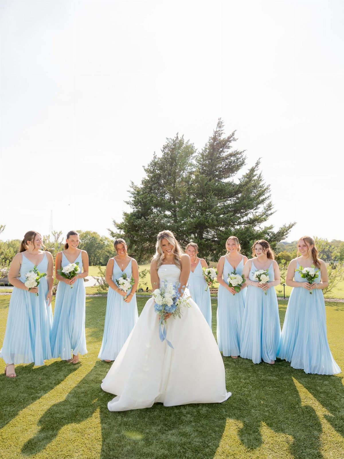 Bride in white gown surrounded by bridesmaids wearing light blue dresses on grass