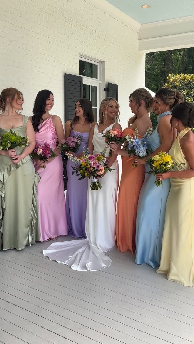 Bride and bridesmaids wearing mismatched colorful dresses holding bouquets on a white porch