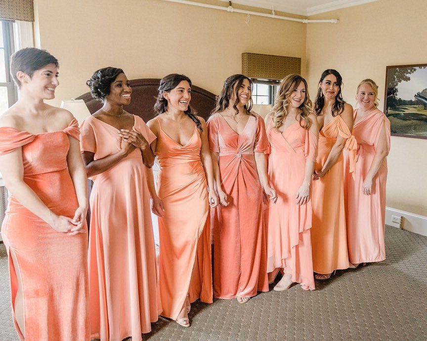 Seven bridesmaids wearing various styles of coral-colored gowns standing in a bedroom.
