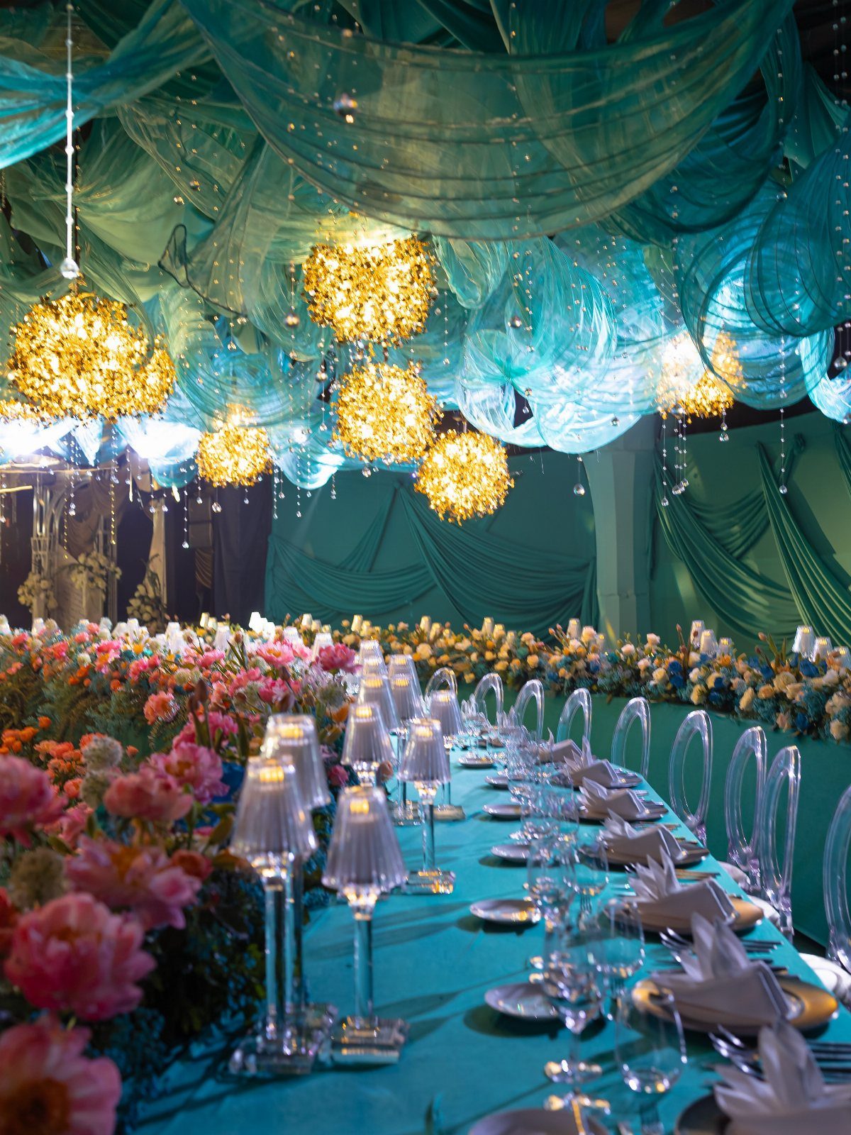 Teal draped event space with gold chandeliers and coral pink floral table arrangements.
