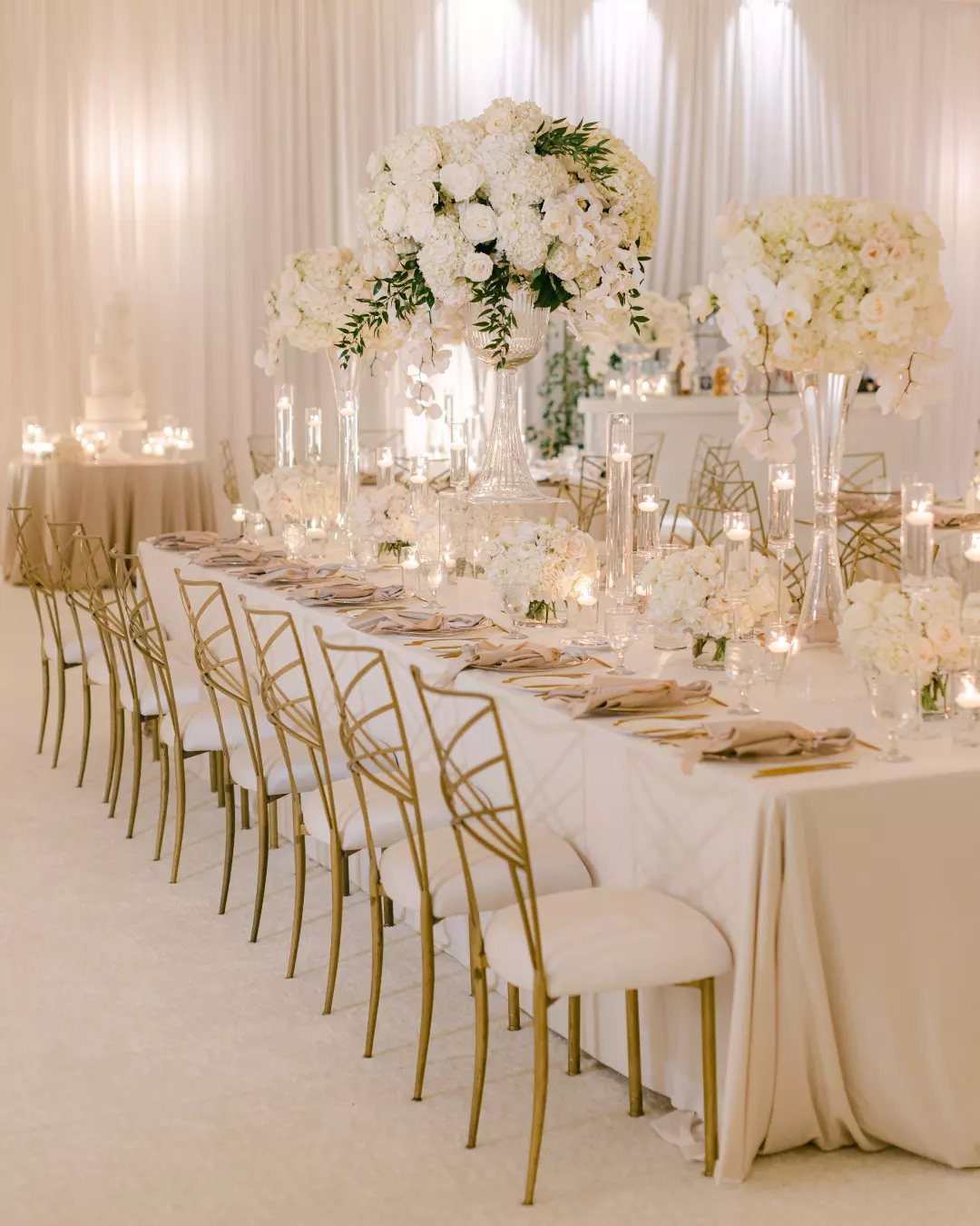 Ivory banquet table with gold chairs and white floral centerpieces in a ballroom.