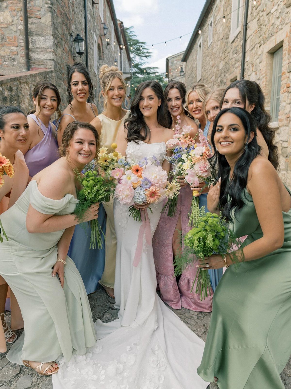 Bride and bridesmaids in multicolored pastel dresses posing in a rustic stone alleyway.