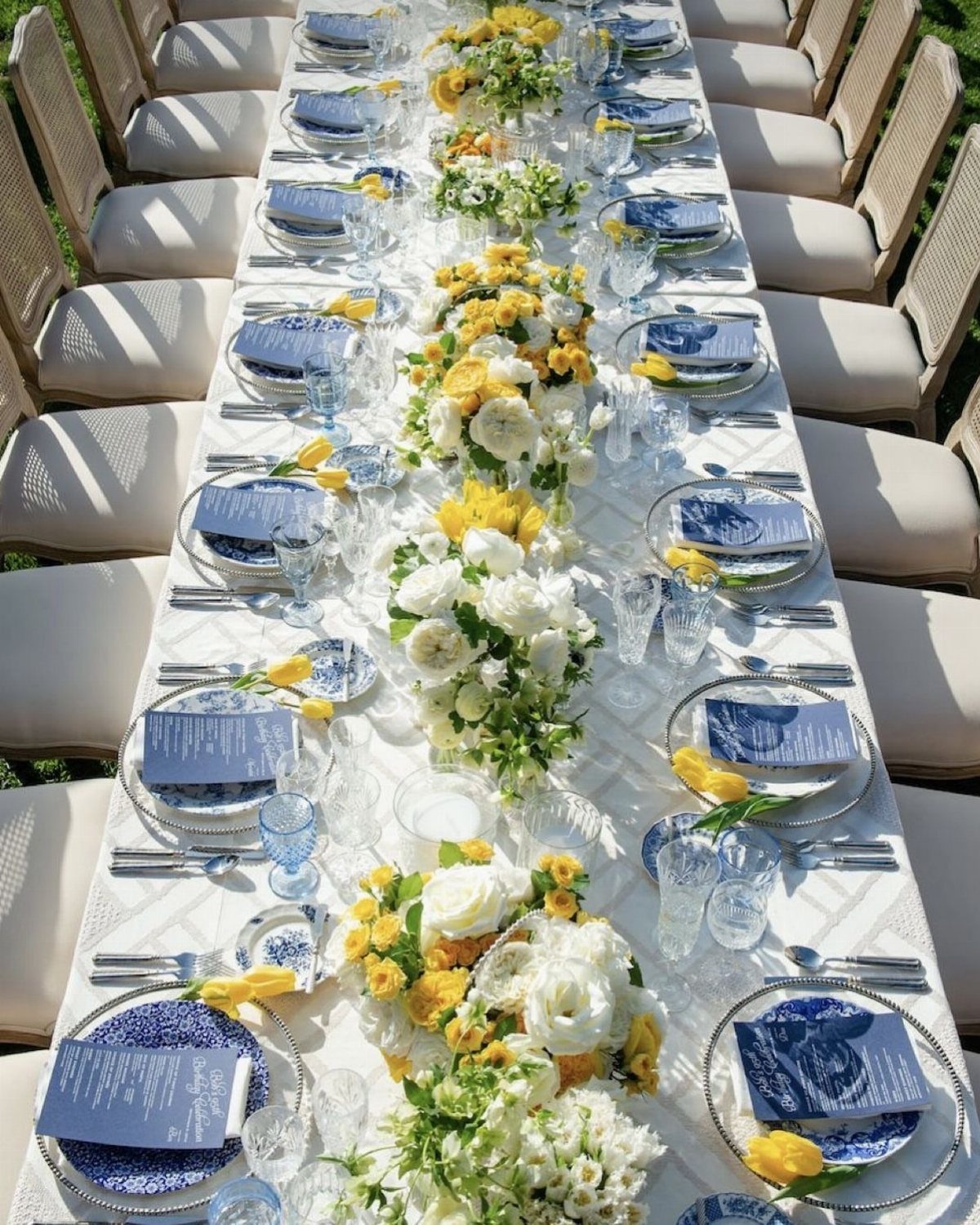 Long dining table with navy blue place settings and yellow and white floral centerpieces.