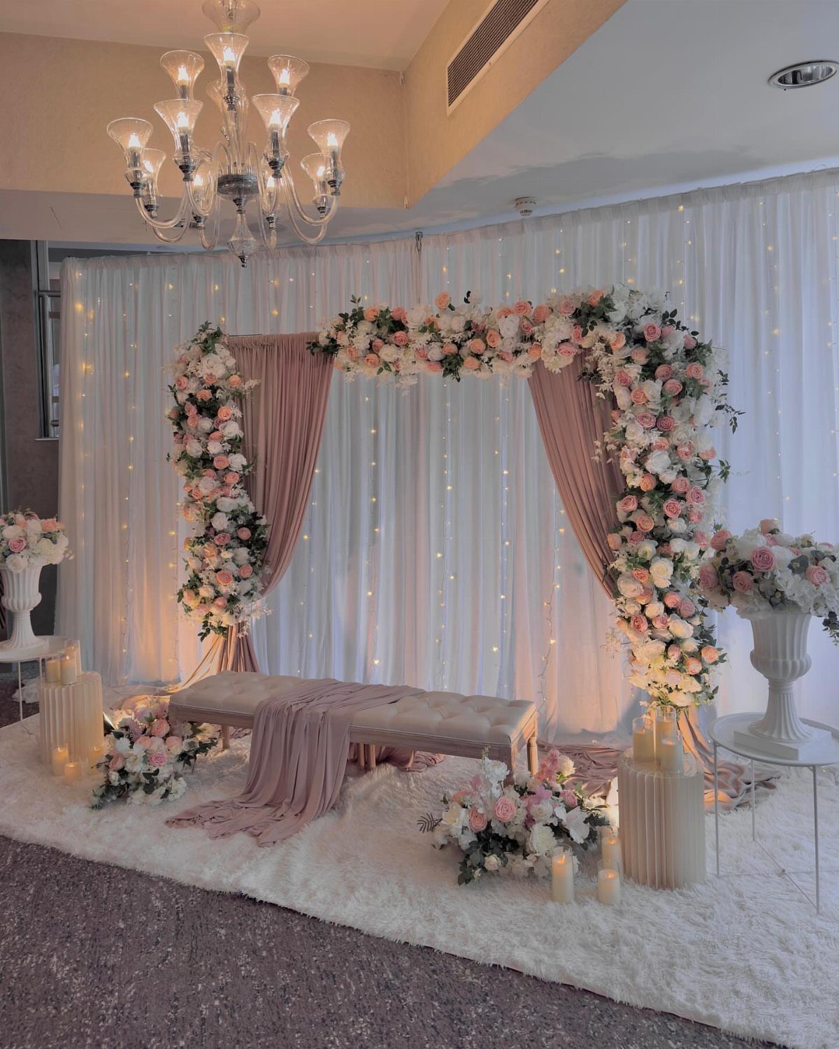 Floral wedding arch with pink and white roses, draped fabric, and crystal chandelier.